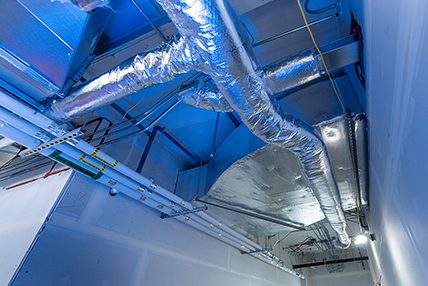 Aluminum-clad ventilation ducts installed on the ceiling of a technical corridor, with bluish lighting and drywall-covered walls