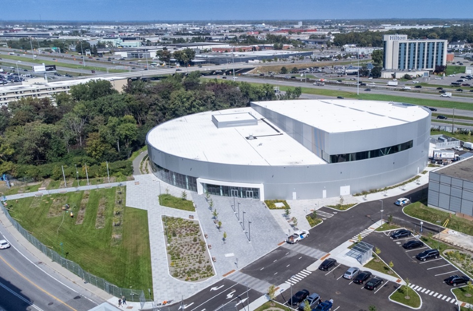 Vue aérienne du Complexe aquatique de Laval, montrant le bâtiment moderne à toit blanc, les aménagements paysagers, le stationnement et l’autoroute en arrière-plan