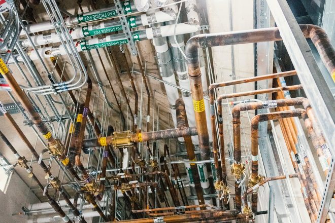 Réseau dense de tuyauterie en cuivre et conduits mécaniques suspendus au plafond d’une salle technique, incluant des étiquettes pour l’air médical et l’eau chaude