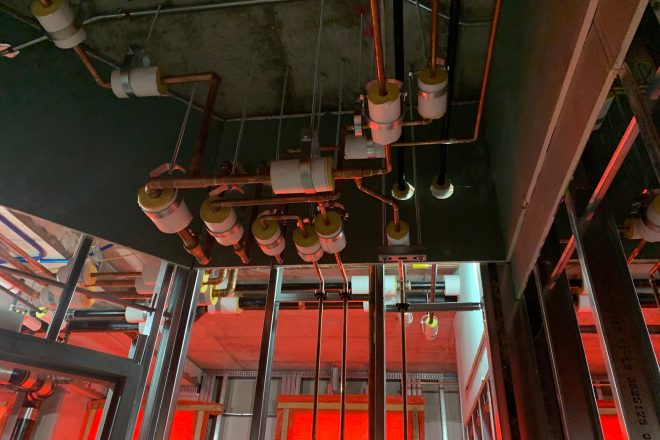 Réseau complexe de conduites en cuivre et en PVC avec isolants suspendus au plafond d’un bâtiment en construction, éclairé par une lumière rouge temporaire