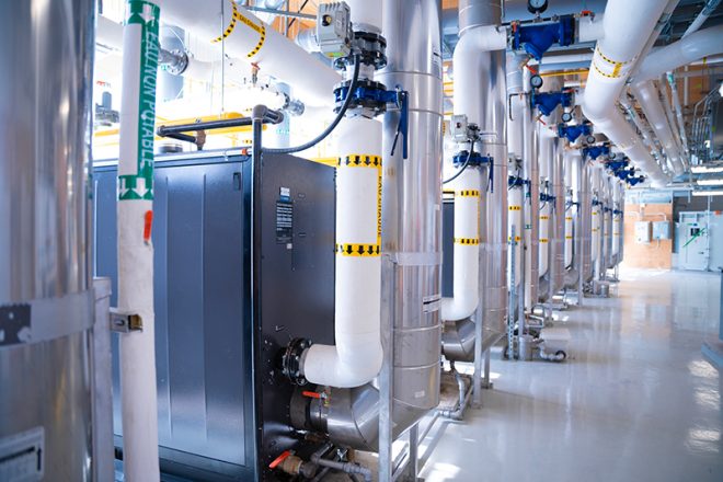 Bright mechanical room with a row of insulated pipes for chilled and hot water, motorized valves, and stainless-steel industrial equipment installed on a polished floor