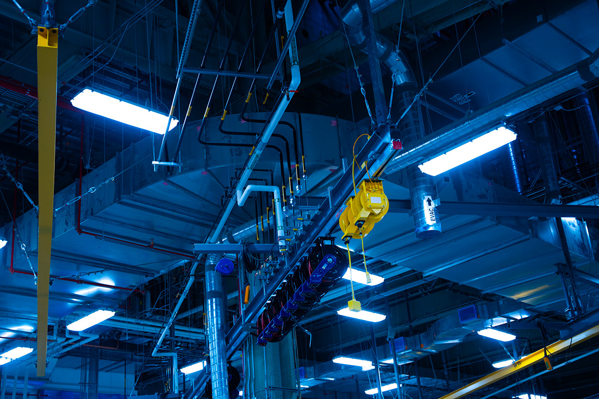 Ceiling of an industrial mechanical room with labeled piping, suspended cable reels, ventilation systems, and bluish fluorescent lighting