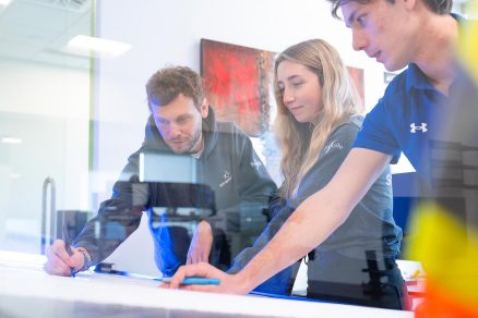 Three young professionals collaborating in a bright, modern office space, analyzing technical plans on a glass table. The image highlights teamwork, innovation, and a dynamic tech work environment.